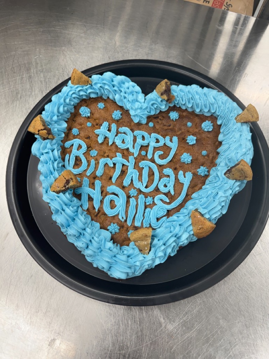 Real heart-shaped chocolate chip cookie cake from BB's Bagels with blue piped frosting border, frosting stars, cookie chunks, and the message Happy Birthday Hailie